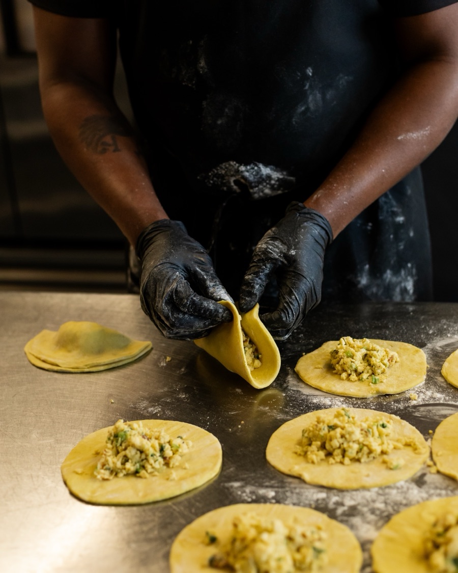 Gloved hands folding a filled round of pattie dough in half, with more filled rounds waiting on the floured counter